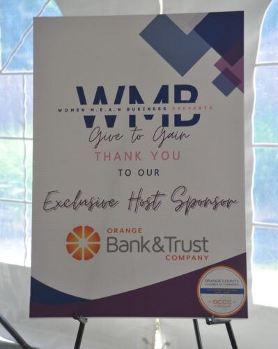 Poster on an easel for Women in M.E.A.N. Business presents 'Give to Gain' with 'Thank You to Our Exclusive Host Sponsor' Orange Bank & Trust Company, featuring orange logo and purple geometric accents.