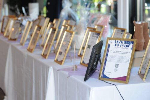 Row of gold-framed certificates lined up on a long white table at an event, with a black digital frame in the foreground.