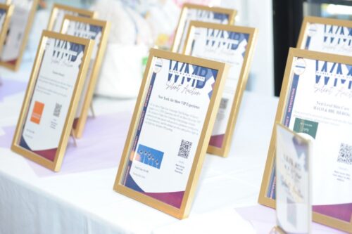 Framed certificates displayed on a white table at an event or expo, arranged in a row with gold frames and purple accents behind them, one foreground frame in focus.