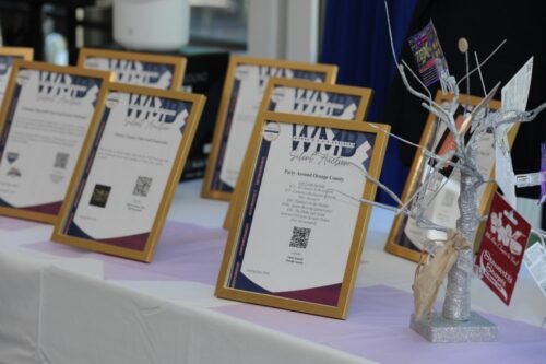 Row of gold-framed certificates displayed on a table at an event, with a silver decorative tree centerpiece on the right.