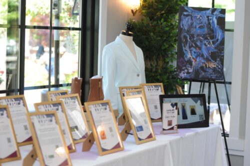 Event display: dress form in a white blazer beside framed certificates, boots, a laptop, and a blue abstract painting on an easel near a window.