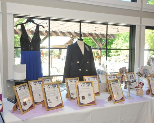 Display table featuring framed certificates beside a blue evening gown and a navy double-breasted blazer on mannequins, with a bright window backdrop.