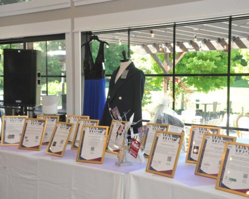 A display table with multiple framed certificates, a black blazer on a mannequin, and a blue-sequin dress in the background.