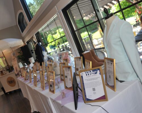 Display table with gold-framed signs and a dress-form wearing a pale blue blazer near large windows; outdoor greenery visible.