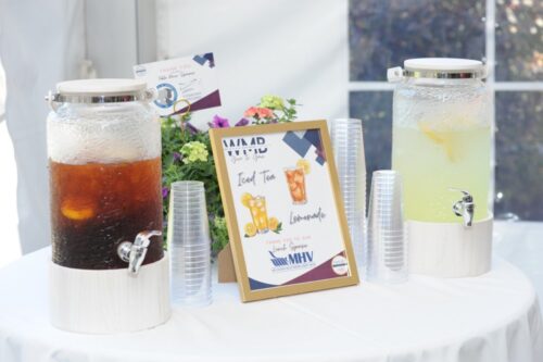 Two large glass beverage dispensers on a white table—iced tea on the left and lemonade on the right—with plastic cups and a framed menu in the center.